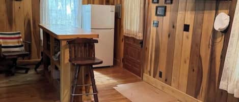 Un interior de fusta amb una zona de cuina, un tamboret, un refrigerador i un televisor penjat a la paret.