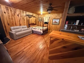 Una habitación con paneles de madera, una cama, un sofá, una mesa de madera y una zona de cocina con un lavamanos.