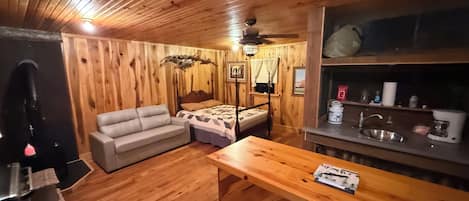 A wooden cabin interior with a bed, a sofa, a dining table, and a kitchenette.