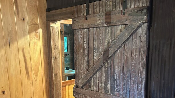 Interior de madera con una puerta de estilo rústico tipo granero, suelo de madera y un baño visible con lavamanos.