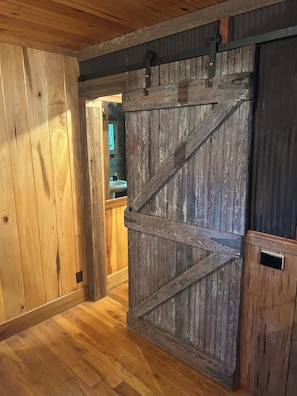 A rustic interior with a wooden floor, a sliding barn-style door, and a visible bathroom with a sink.
