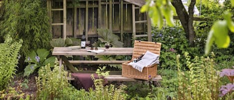 Een houten prieel met een bankje en picknicktafel, omringd door weelderige begroeiing en planten.