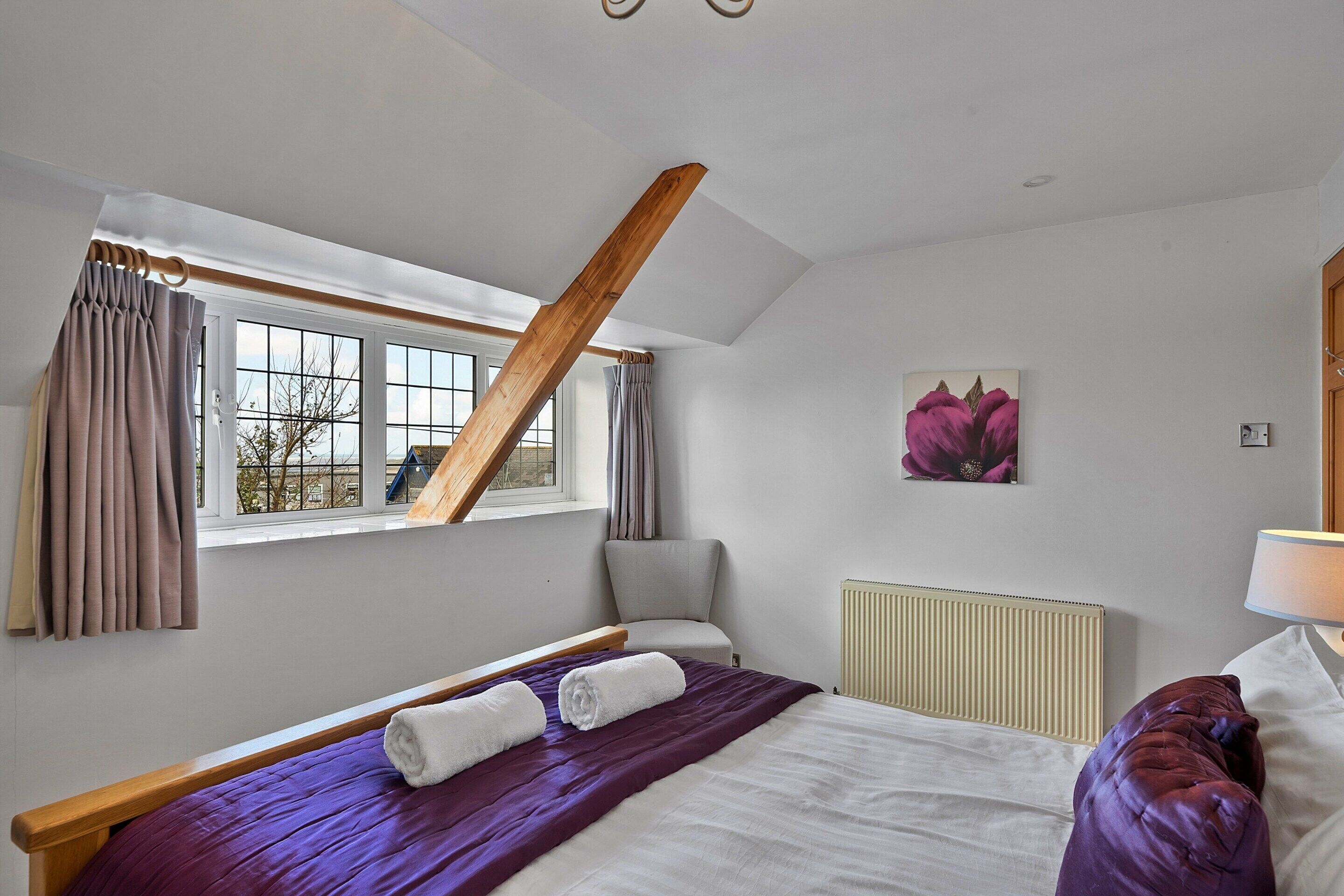 A bedroom with a sloped ceiling, a bed with a purple bedspread, a grey armchair, and a radiator.