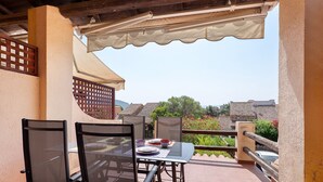 Eine Terrasse im Freien mit Tisch und Stühlen, einem Pergola darüber und Blick auf Gebäude und Grünflächen im Hintergrund.