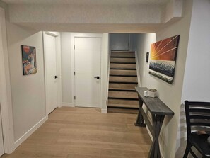 A hallway with a wooden floor, a white door, a staircase, and a dining area with a table and chairs. - Come Stay at "Night at the Movies" with all the comforts of home! (Calgary)