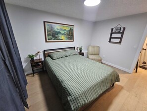 Un dormitorio con cama, una silla, una mesita de noche, una lámpara, un cuadro enmarco y un letrero motivacional.