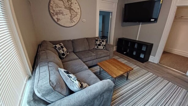 En stue med en grå sofa, et træbord, et verdenskort på væggen, et fjernsyn og en dør, der fører til et andet rum.