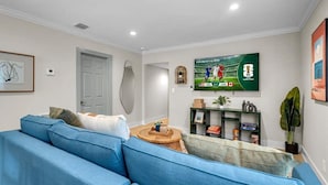 Een woonkamer met een blauwe bank, een televisie waarop de FIFA Wereldbeker te zien is, en een deur op de achtergrond.