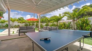 Een pingpongtafel buiten, met een plafondventilator, omringd door buitenmeubels en palmbomen.