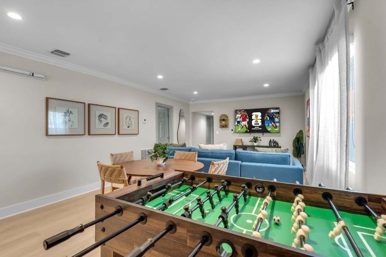 A room with a foosball table, a blue sofa, a dining table with chairs, and framed artwork on the wall.