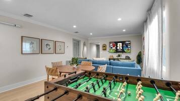 A room with a foosball table, a blue sofa, a dining table with chairs, and framed artwork on the wall.