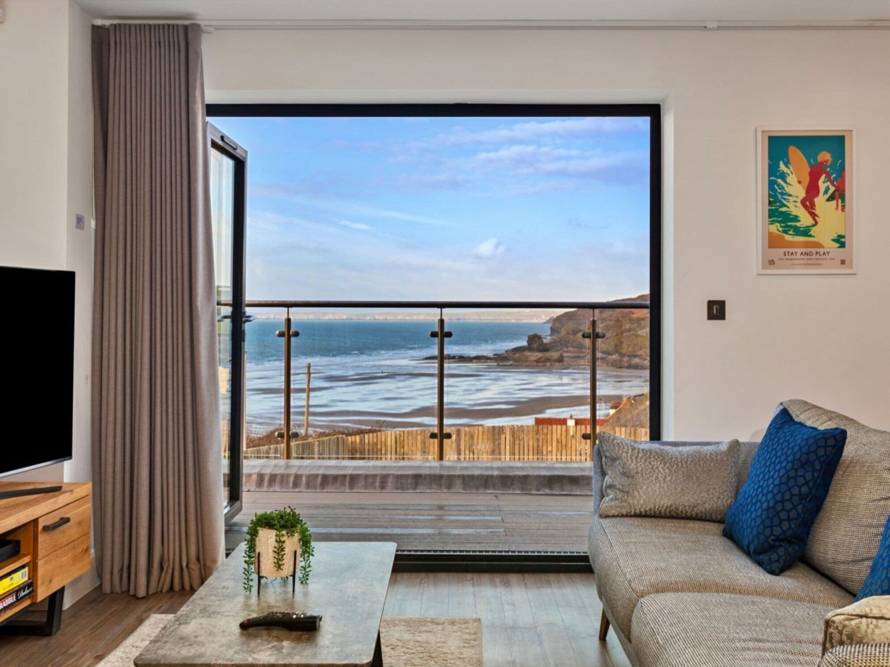 A living room with a large window overlooking a beach, a sofa, a coffee table, and a television.