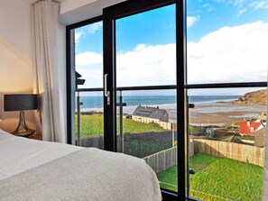 A bedroom with a large window overlooking a beach and a view of a house across the water. - Stunning Broad Haven Beach House (Haverfordwest)