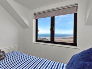 Ein Schlafzimmer mit großem Fenster und Blick auf einen Strand, einem Bett mit gestreifter Bettwäsche und einem Heizkörper.
