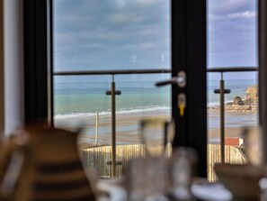 Blick durch ein Fenster mit Metallgitter auf einen Strand mit Wellen und felsigen Klippen.