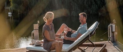 A couple sitting on a wooden lounge chair by a water body, with a wooden deck and a small table with a hat and glass.