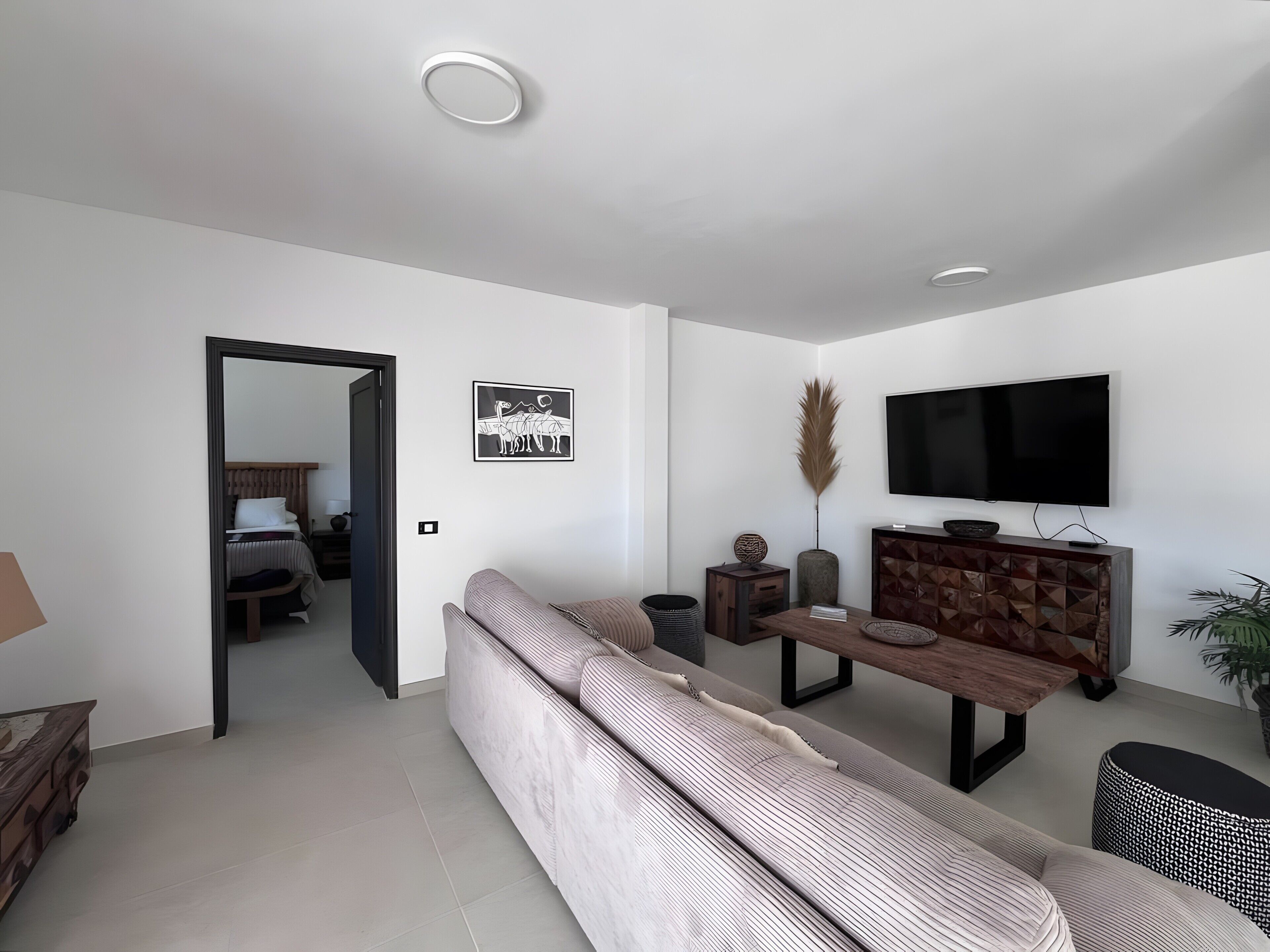 A modern living room with a sofa, a dining table, a TV mounted on the wall, and a door leading to another room.