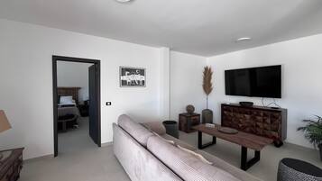 A modern living room with a sofa, a dining table, a TV mounted on the wall, and a door leading to another room.