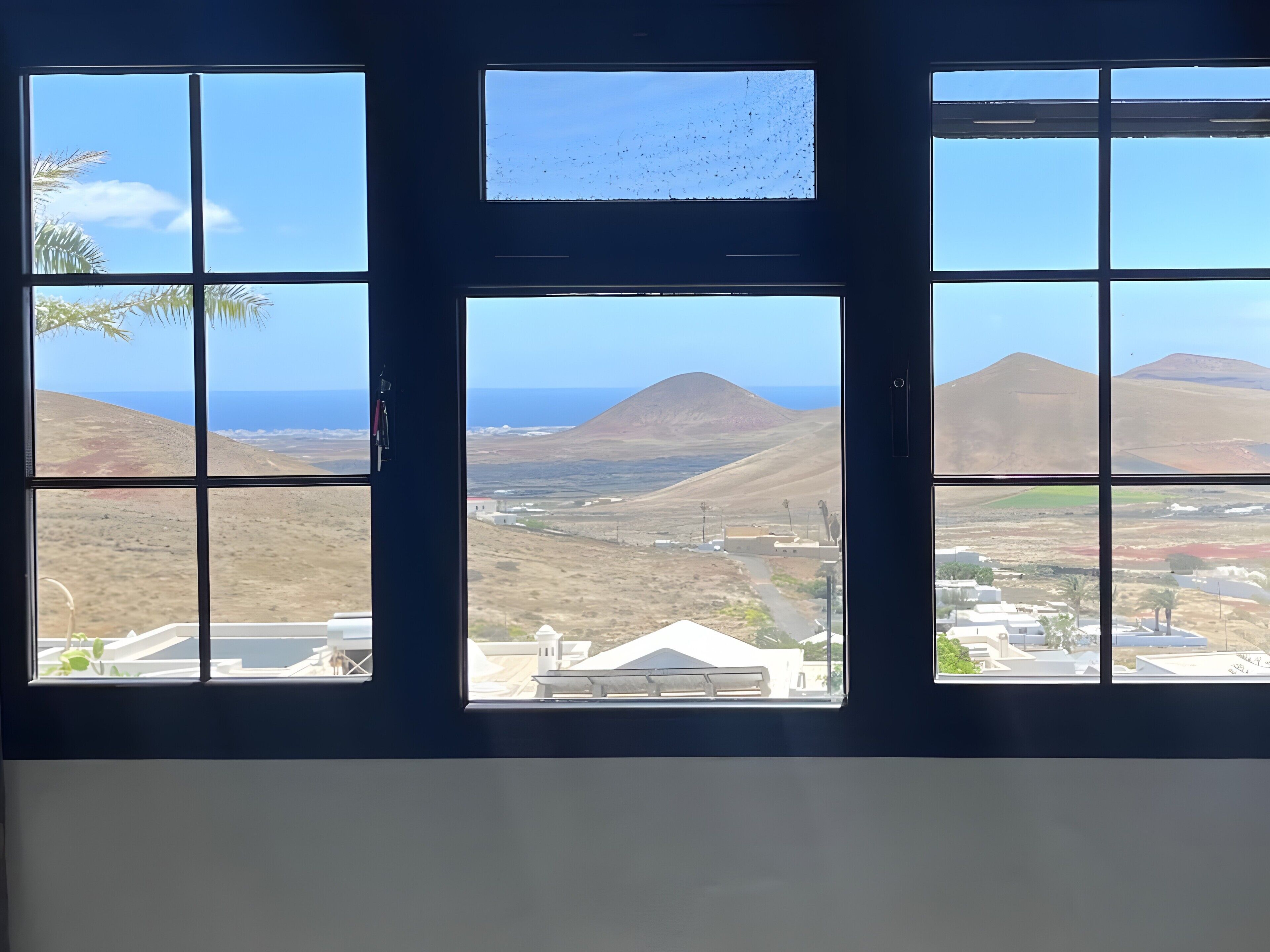 Une pièce dotée d’une grande fenêtre divisée en plusieurs panneaux, qui offre une vue sur un paysage côtier avec des collines et un ciel dégagé.