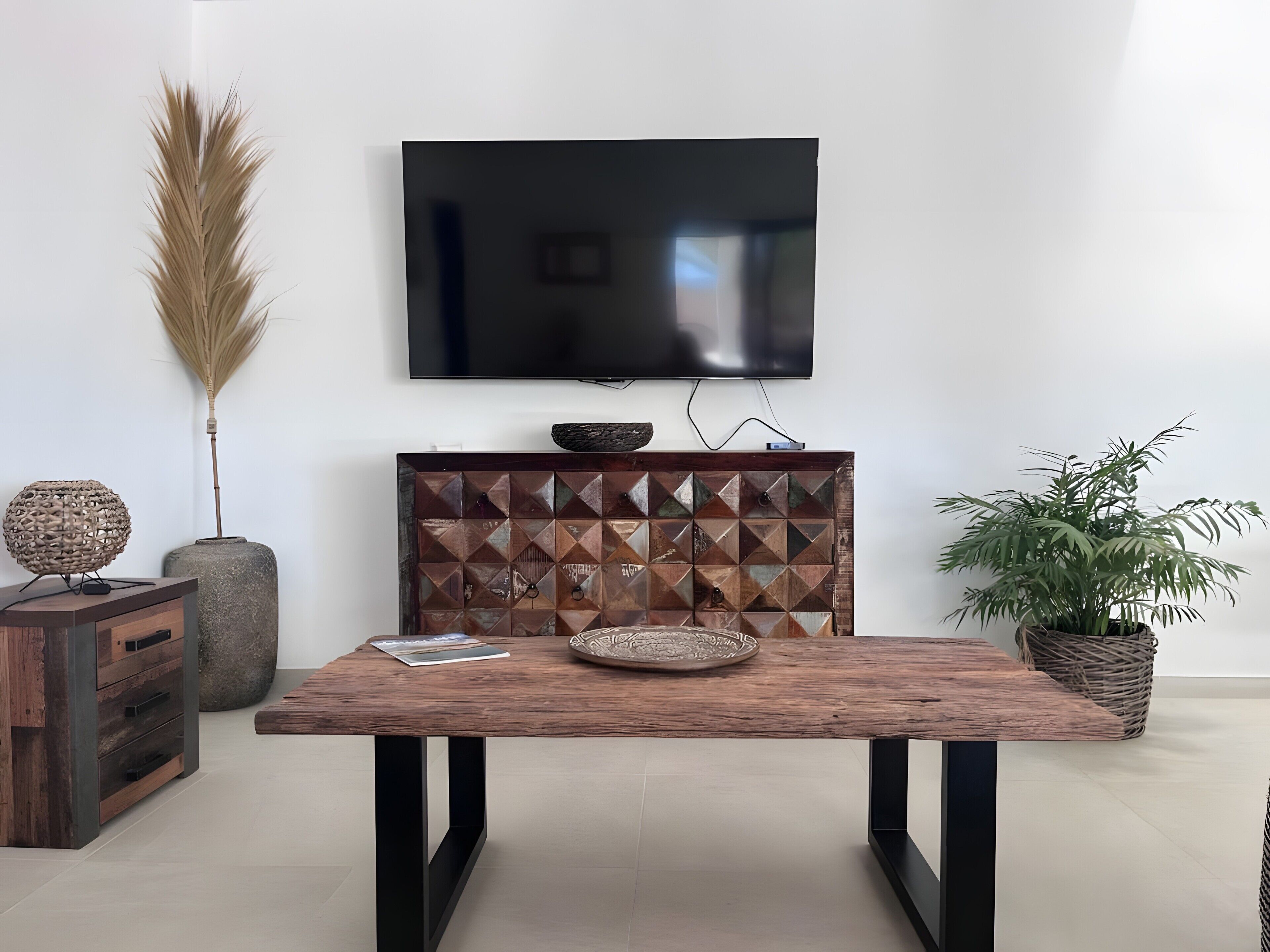 A modern living room with a wooden coffee table, a flat-screen TV mounted on the wall, a decorative cabinet, and a potted plant.