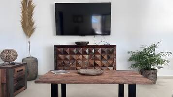 A modern living room with a wooden coffee table, a flat-screen TV mounted on the wall, a decorative cabinet, and a potted plant.