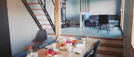 A dining area with a wooden table set for breakfast, a staircase, and a modern living space in the background.