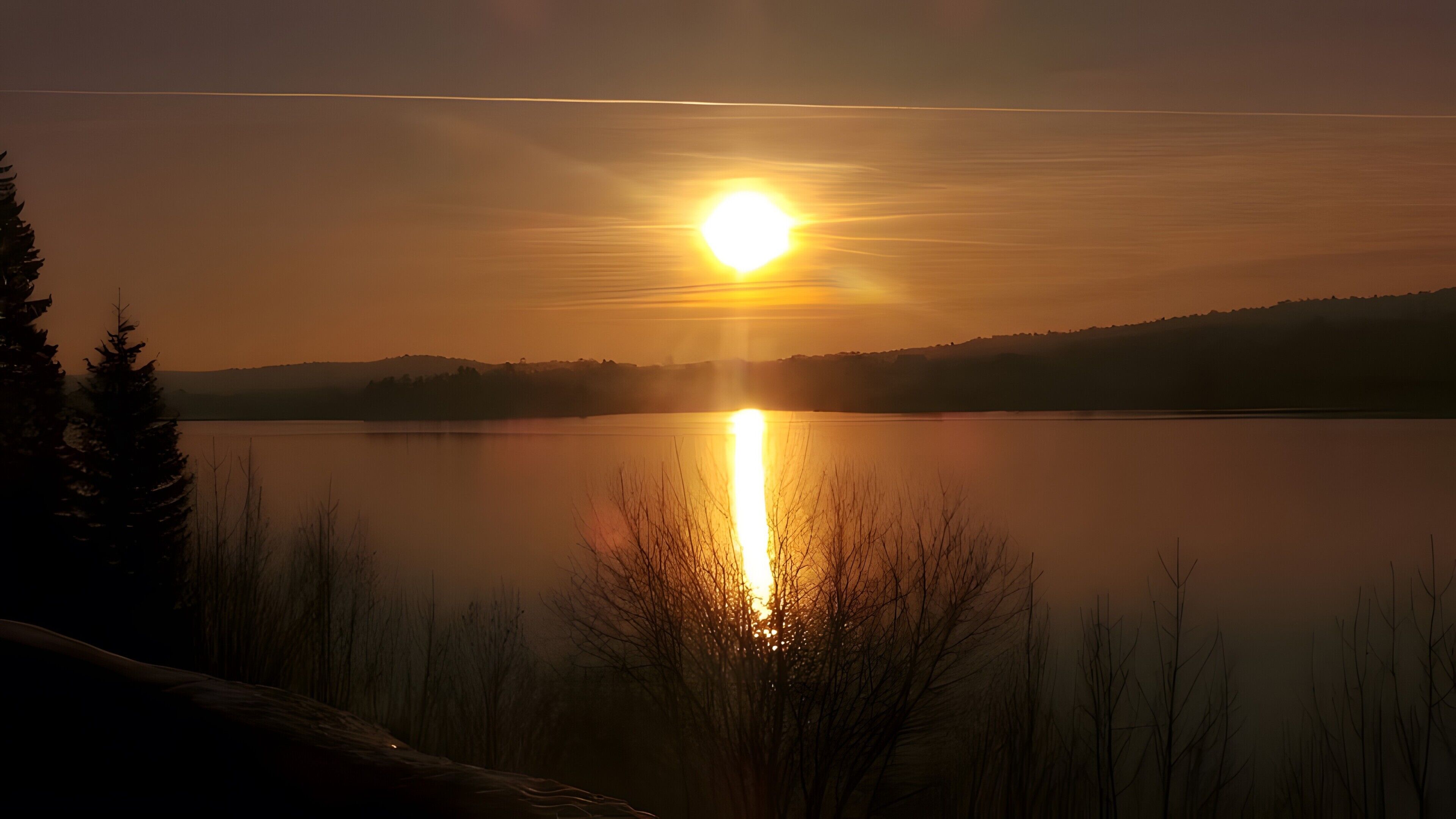A sunset over a calm lake with a silhouette of trees and hills in the background.