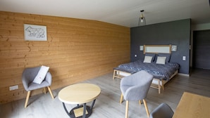 A modern bedroom with a wooden wall, a bed with patterned bedding, two grey armchairs, a round wooden table, and a hanging light fixture. - Bed and Breakfast 'Chambre La Grande Ourse' with Lake View, Shared Terrace and Wi-Fi (Grande-Rivière Château)