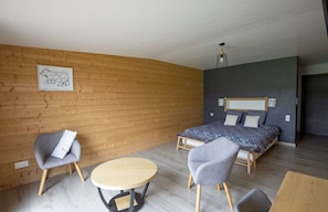 A modern bedroom with a wooden wall, a bed with patterned bedding, two grey armchairs, a round wooden table, and a hanging light fixture. - Bed and Breakfast 'Chambre La Grande Ourse' with Lake View, Shared Terrace and Wi-Fi (Grande-Rivière Château)
