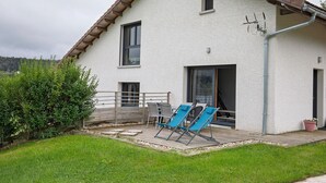 Una casa con fachada blanca, techo marrón y una ventana pequeña. Hay un patio con dos sillas azules y una mesa.