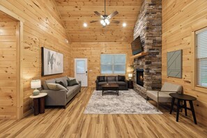 A cozy cabin interior with a fireplace, wooden walls, a ceiling fan, and a large window.