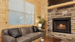 A cozy living room with a stone fireplace, a leather sofa, and a wooden coffee table. - Dollys Cabins 1010  - Walk to Dollywood! (Pigeon Forge)