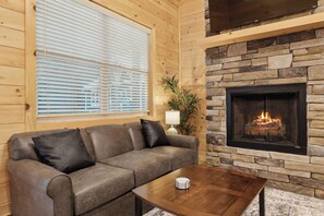 A cozy living room with a stone fireplace, a leather sofa, and a wooden coffee table. - Dollys Cabins 1010  - Walk to Dollywood! (Pigeon Forge)