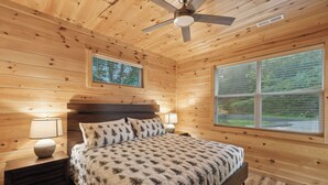 A wooden cabin bedroom with a bed, two bedside lamps, a ceiling fan, and two windows. - Dollys Cabins 1010  - Walk to Dollywood! (Pigeon Forge)
