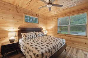 A wooden cabin bedroom with a bed, two bedside lamps, a ceiling fan, and two windows. - Dollys Cabins 1010  - Walk to Dollywood! (Pigeon Forge)
