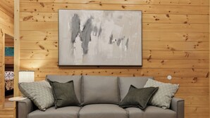 A room with a grey leather sofa against a wooden wall, a framed abstract painting above it, and a small table with a lamp to the left. - Dollys Cabins 1010  - Walk to Dollywood! (Pigeon Forge)
