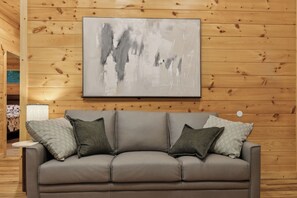 A room with a grey leather sofa against a wooden wall, a framed abstract painting above it, and a small table with a lamp to the left. - Dollys Cabins 1010  - Walk to Dollywood! (Pigeon Forge)