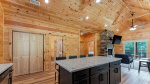 A wooden cabin interior with a kitchen area featuring a granite countertop, a stone fireplace, and a dining area with outdoor seating. - Dollys Cabin 1012  - Walk to Dollywood! (Pigeon Forge)