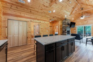 Ein Holzhütten-Interieur mit Küchenbereich, der eine Granitarbeitsplatte, einen Kamin aus Naturstein und eine Essecke mit Außensitzmöbeln zeigt.