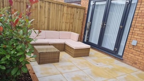 Eine Terrasse mit einer Eckcouch und zwei Hocker, einem Holzzaun und einer Glastür.