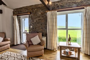 Una habitación acogedora con pared de piedra, un ventanal con cortinas, una silla de madera, una mesita con libros y una alfombra estampada.