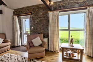 Una habitación acogedora con pared de piedra, un ventanal con cortinas, una silla de madera, una mesita con libros y una alfombra estampada.