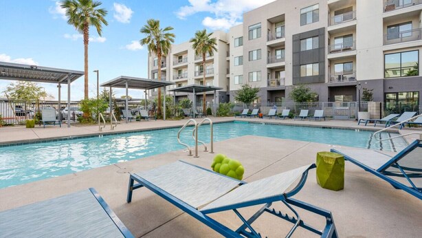 A swimming pool with lounge chairs and umbrellas, surrounded by a multi-story building with balconies. - 2BR Desert Getaway - Pool, King Beds, Hot Tub, Gym (Tempe)