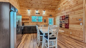 A cabin-style kitchen with a central island, wooden walls, and a dining area with white chairs. - Dollys Cabins 1014 - Walk to Dollywood! (Pigeon)