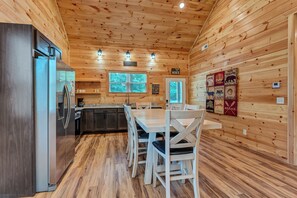 A cabin-style kitchen with a central island, wooden walls, and a dining area with white chairs. - Dollys Cabins 1014 - Walk to Dollywood! (Pigeon)