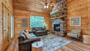 A cozy cabin interior with a stone fireplace, wooden walls, a ceiling fan, and a large window. - Dollys Cabins 1014 - Walk to Dollywood! (Pigeon)