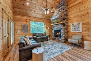 A cozy cabin interior with a stone fireplace, wooden walls, a ceiling fan, and a large window. - Dollys Cabins 1014 - Walk to Dollywood! (Pigeon)