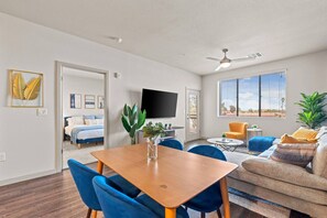 A modern living room with a dining area, a flat-screen TV mounted on the wall, and a large window with blinds. - 2BR Tempe Oasis - King Beds, Pool, Gym, DT Tempe (Tempe)