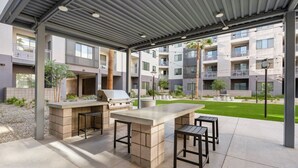 Eine Terrasse im Freien mit Grill und Sitzgelegenheit, umgeben von einer überdachten Struktur und Blick auf Wohnhäuser.