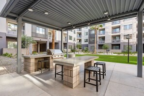 Eine Terrasse im Freien mit Grill und Sitzgelegenheit, umgeben von einer überdachten Struktur und Blick auf Wohnhäuser.